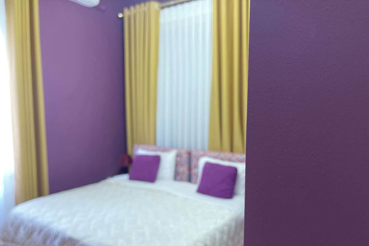 Un lit recouvert d’une couverture blanche et orné de deux coussins violets, une fenêtre avec des rideaux jaunes et un mur violet.