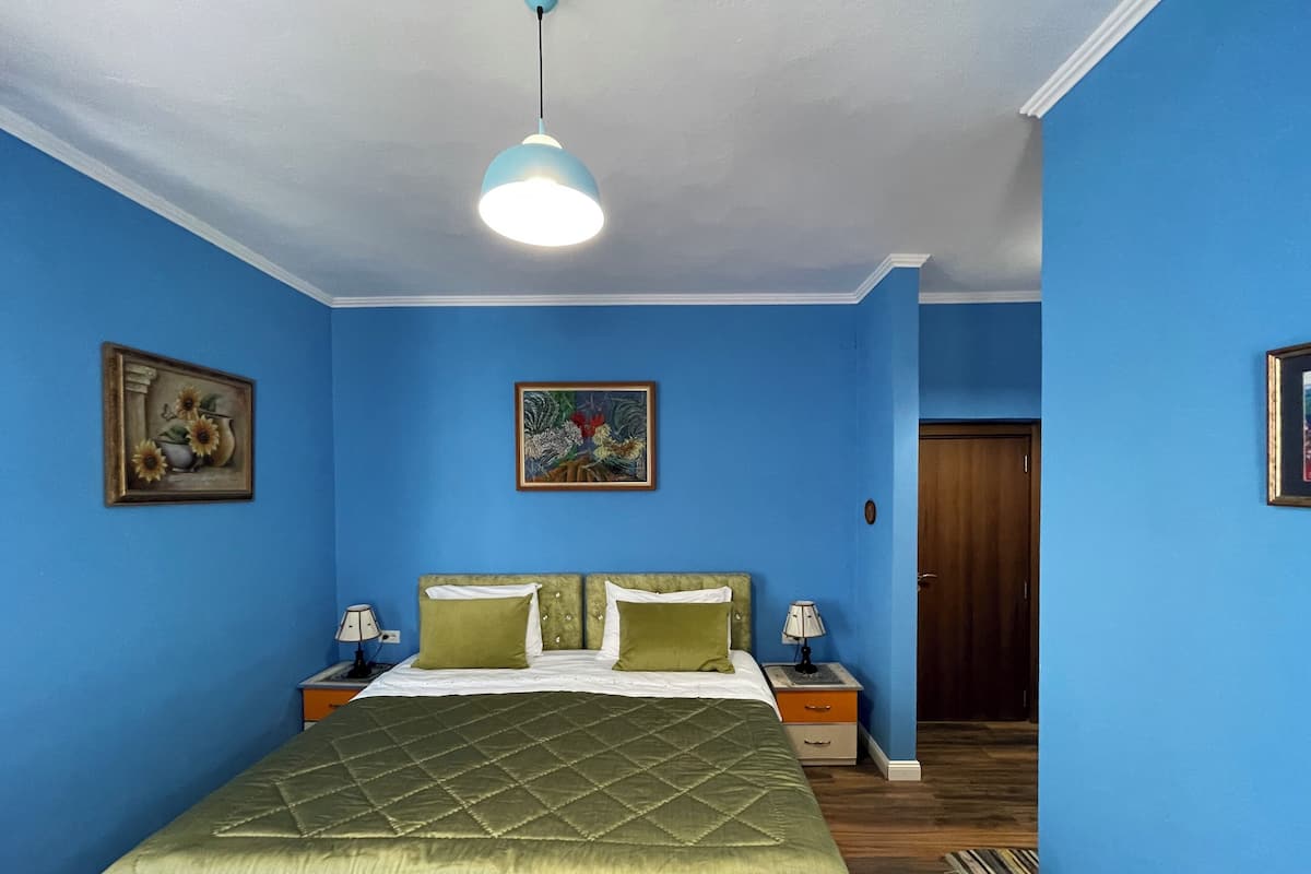Une chambre à coucher comprenant un lit, deux tables de chevet, un luminaire au plafond et deux tableaux accrochés au mur.