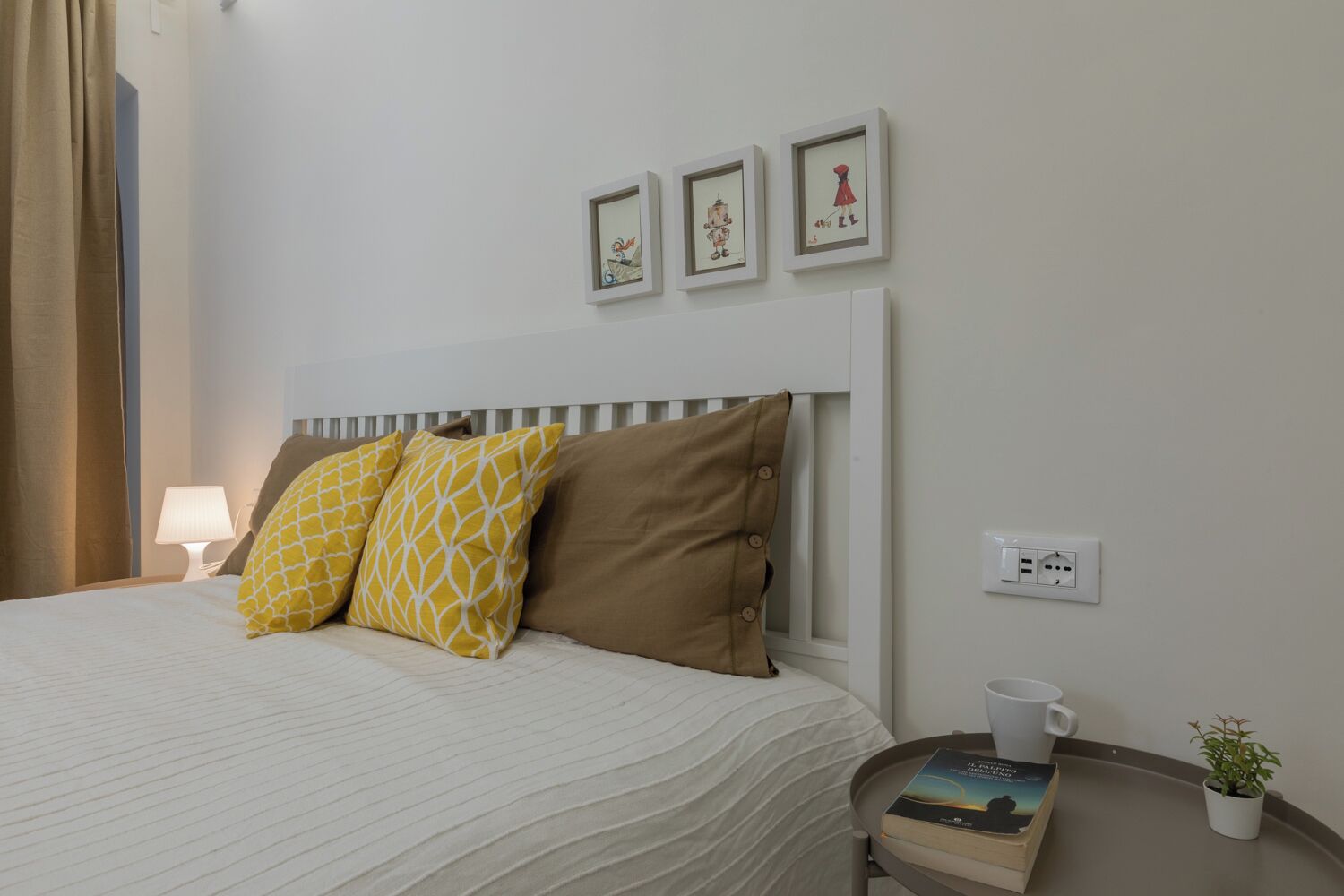 A neatly made bed with patterned pillows, a bedside table with a book and a mug, a small plant, and a wall-mounted power outlet.