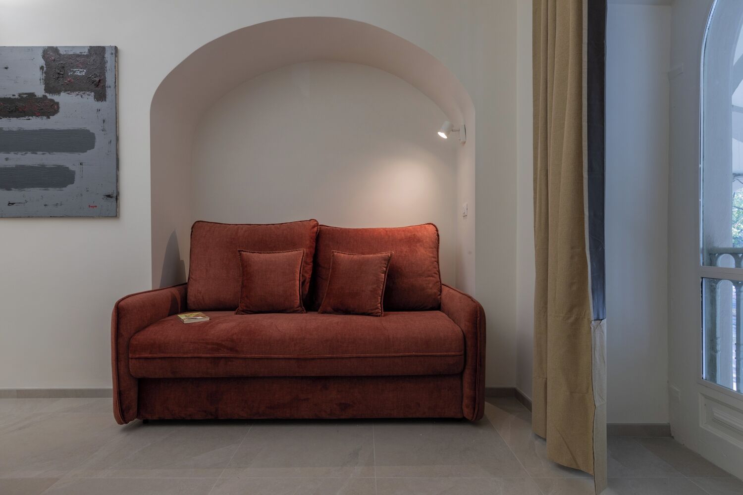 Un canapé en velours rouge avec deux coussins, dans une pièce dont le mur est blanc, ornée d’un tableau et dotée d’une fenêtre avec des rideaux.