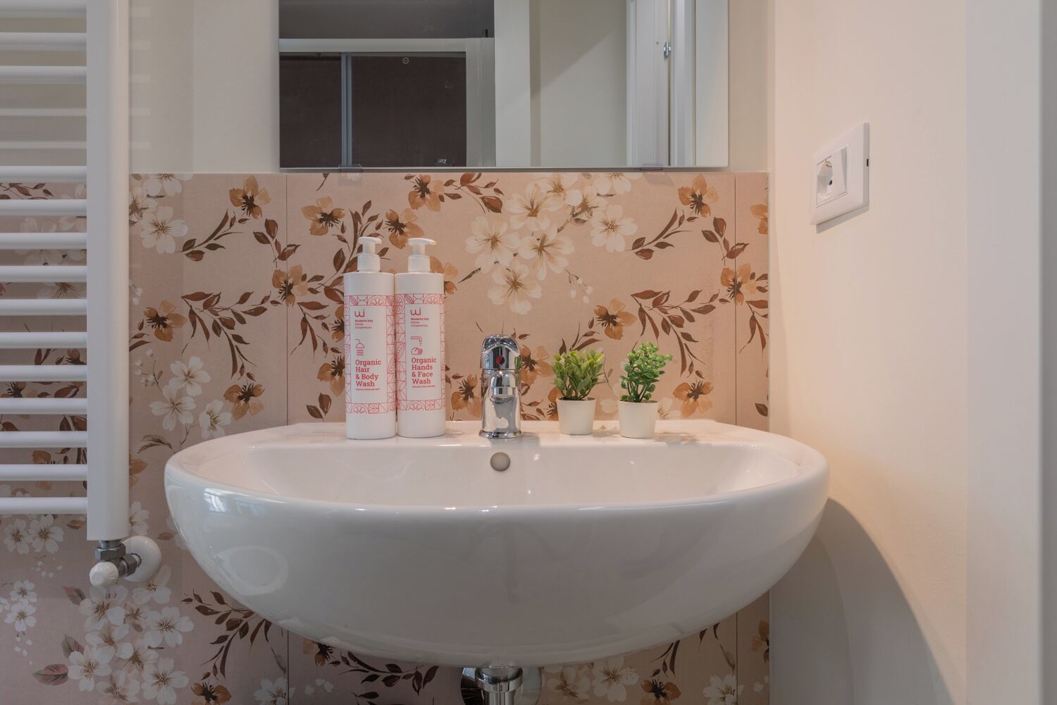 A bathroom with a floral wallpaper, a white sink, two bottles of organic hair and body wash, a small potted plant, and a window above the mirror.