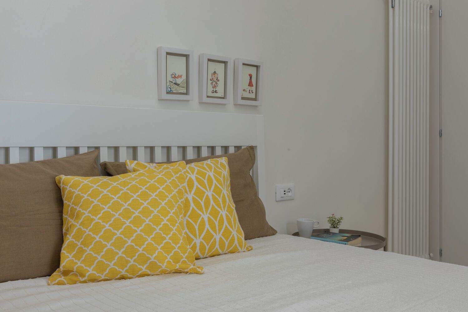 A neatly made bed with a white and brown headboard, two yellow pillows with a white pattern, and a small bedside table with a cup and a plant.