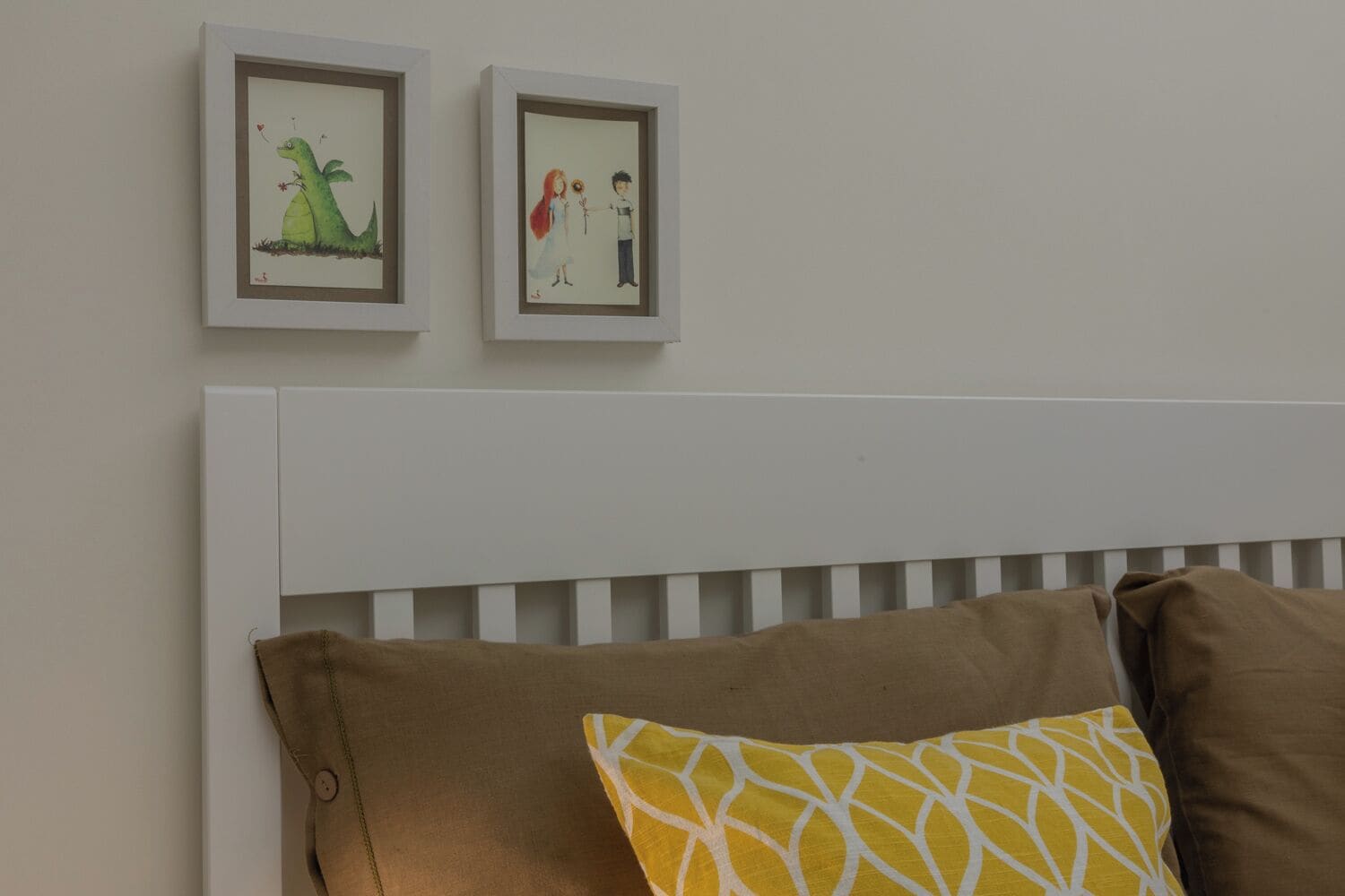 A bedroom with a white headboard, two framed pictures on the wall, and three pillows on the bed.