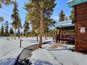 Una cabaña de madera con un porche cubierto, rodeada de suelo cubierto de nieve y altos pinos.