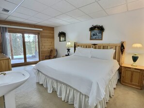 Une chambre à coucher avec un grand lit, une tête de lit en bois, une table de chevet avec une lampe, une chaise en bois, une fenêtre donnant sur un beau paysage et une salle de bain avec un lavabo.
