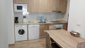 A compact kitchen with a microwave, washing machine, and dishwasher. There is a wooden table and a basket on the counter. - HOSTAL LOS VALLES  (Bielsa)