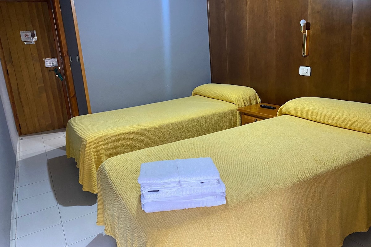 A hotel room with two single beds, a wooden door, a small table, and a wall socket.