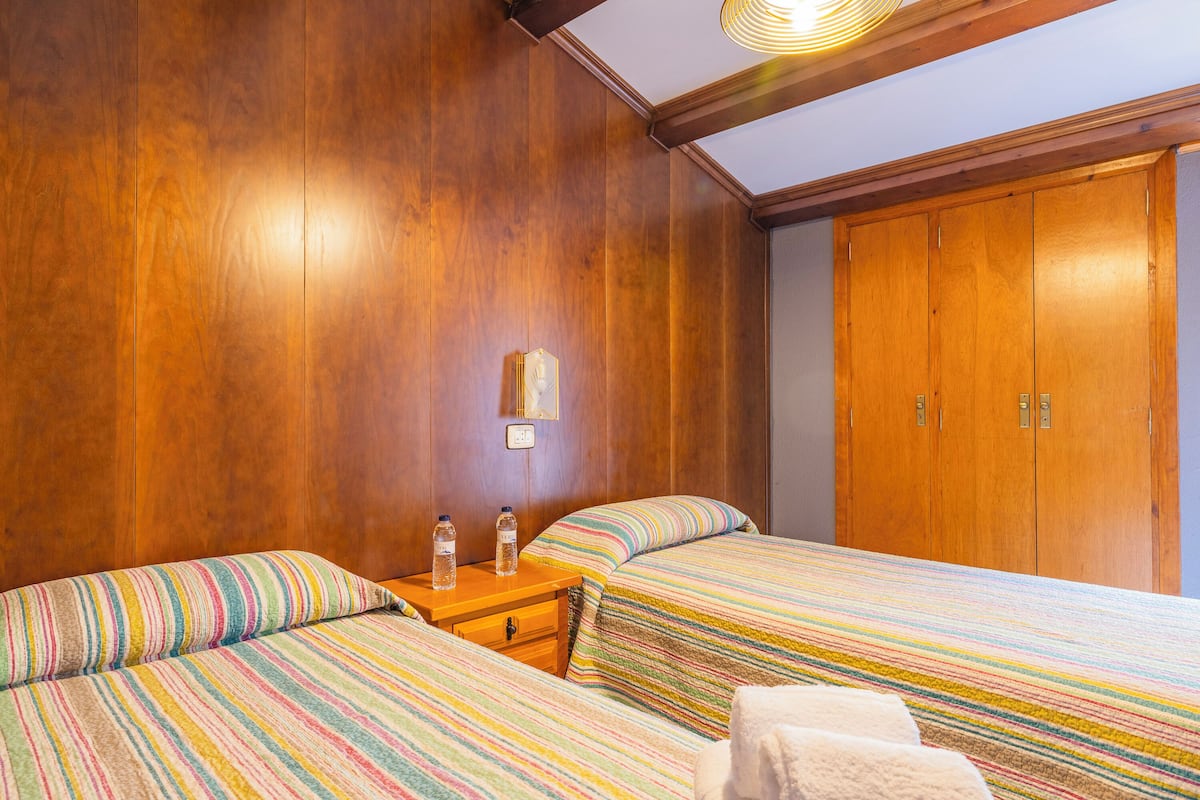 A hotel room with wooden panel walls, a wooden wardrobe, two beds with striped bedding, a nightstand with two water bottles, and a wall-mounted light fixture.