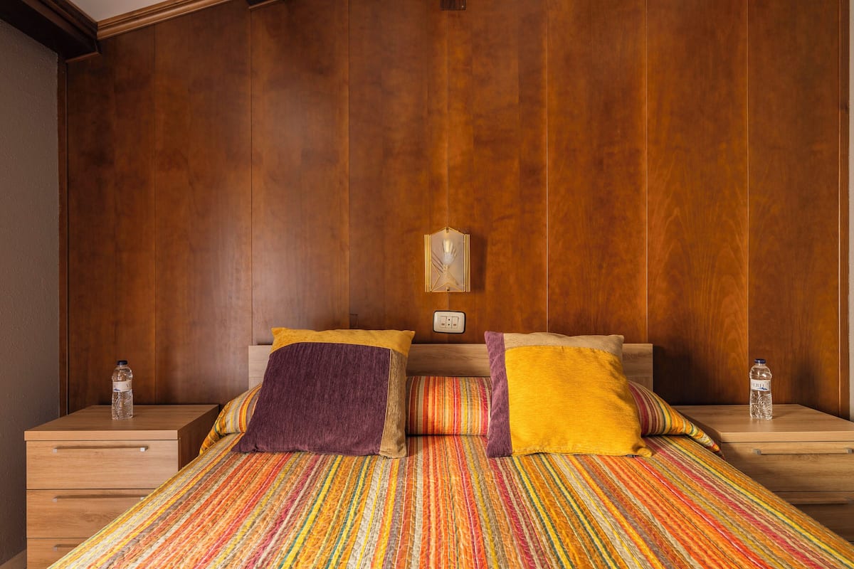 A bedroom with a wooden headboard, a striped bedspread, and two bedside tables with water bottles.