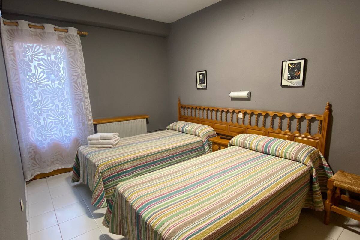 A bedroom with two single beds, a wooden headboard, a nightstand, a window with a curtain, and a ceiling light.
