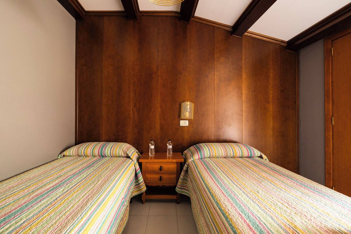 A room with two beds, wooden walls, a small wooden nightstand with two bottles, and a wall-mounted light.