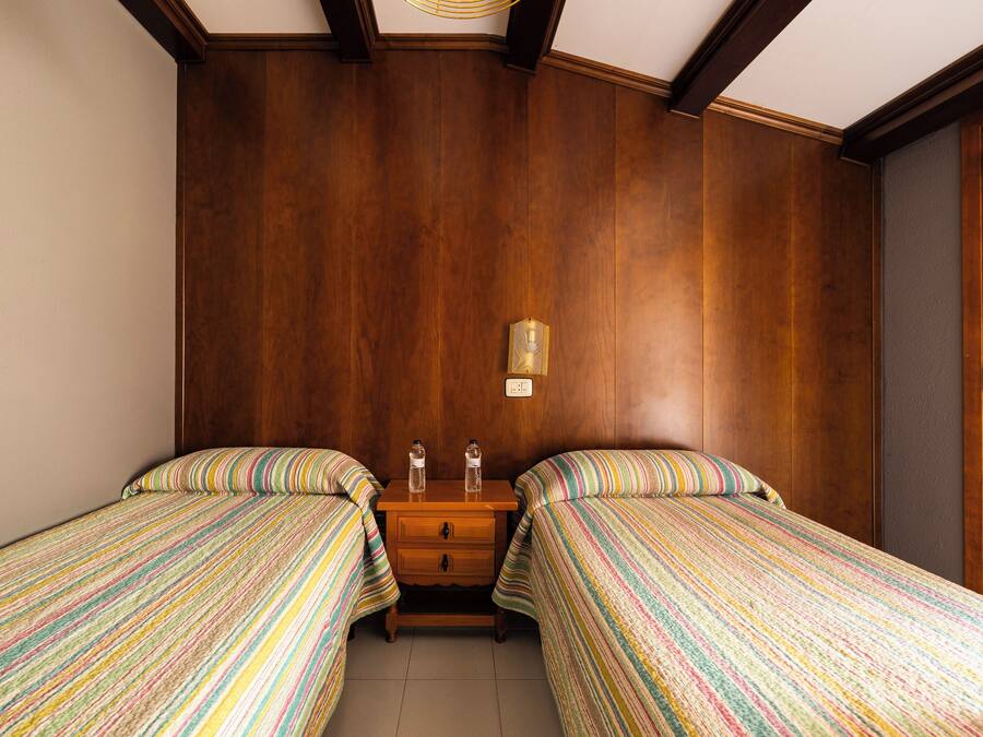 A room with two beds, wooden walls, a small wooden nightstand with two bottles, and a wall-mounted light.