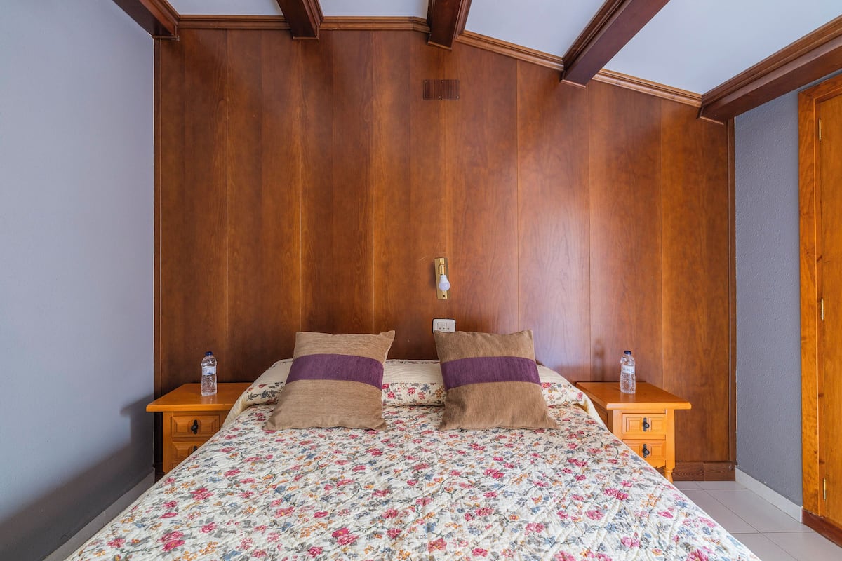 A bedroom with a wooden headboard, floral bedspread, two pillows, a nightstand with a water bottle, and a wooden dresser.