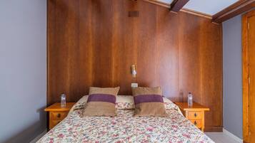 Une chambre à coucher avec une tête de lit en bois, une couvre-lit à motifs floraux, deux oreillers, une table de chevet avec une bouteille d’eau et une commode en bois.