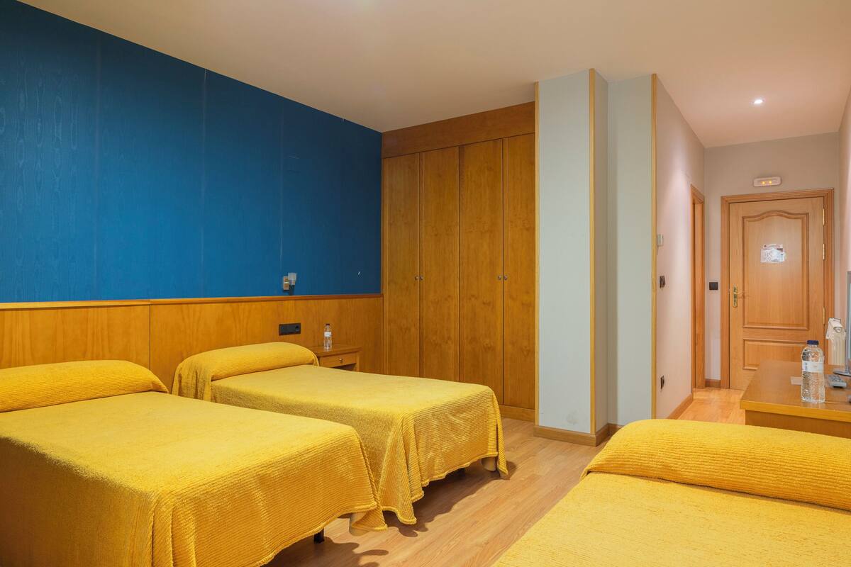 A hotel room with two beds, a wooden wardrobe, a small table with a bottle, and a blue wall.