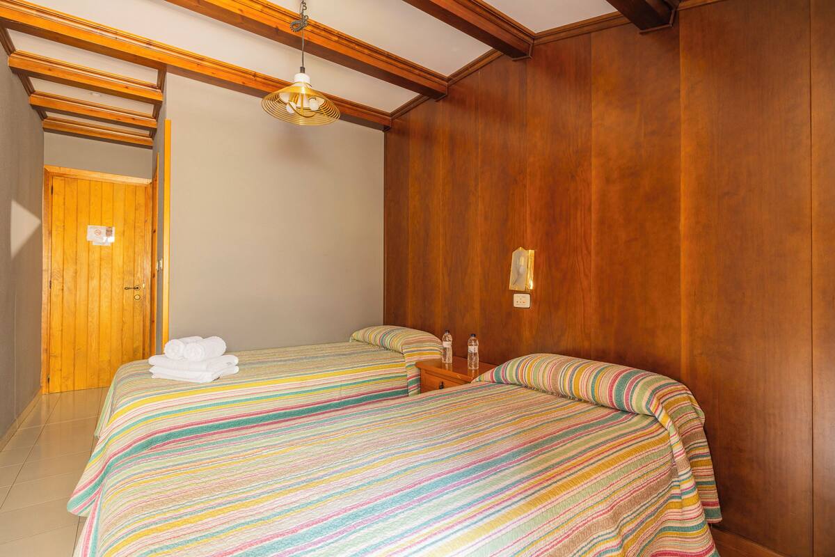 A hotel room with two beds, wooden walls, a wooden door, a hanging light fixture, and a small bedside table with a bottle and a glass.