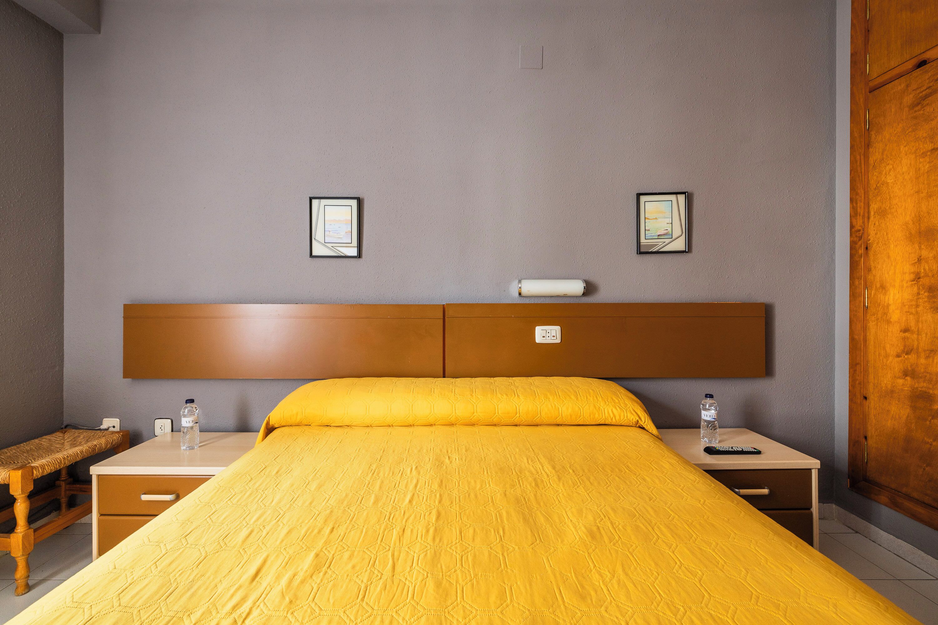 Una habitación de hotel con una cama, mesitas de noche, un banco y dos cuadros en la pared.