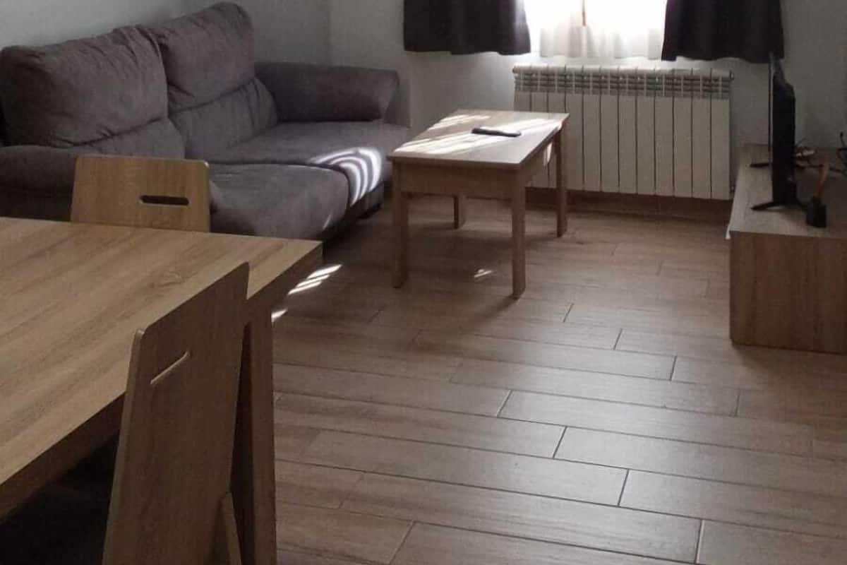 A living room with a sofa, a wooden table, and a coffee table. There is a window with curtains and a radiator.