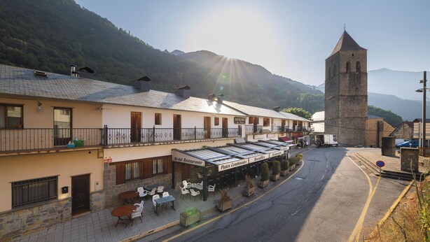 A hotel with outdoor seating, a bell tower, and a mountain backdrop. - HOSTAL LOS VALLES  (Bielsa)