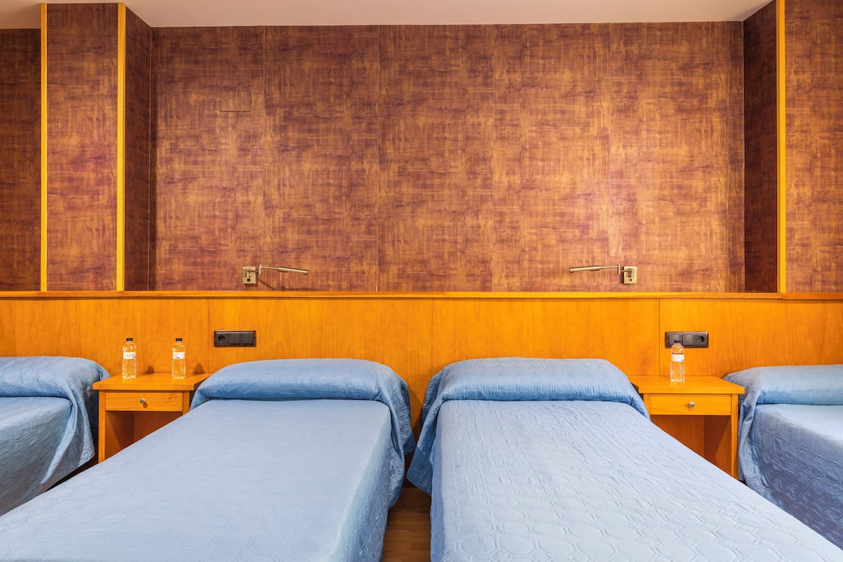 A hotel room with four single beds, wooden headboards, and bedside tables with water bottles.