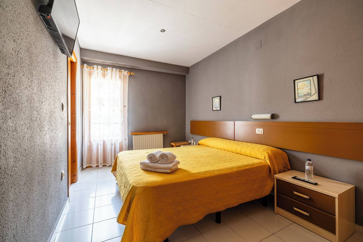A hotel room with a bed, a bedside table, a nightstand, a window with curtains, and a wall-mounted picture.
