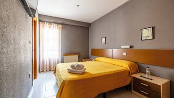 A hotel room with a bed, a bedside table, a nightstand, a window with curtains, and a wall-mounted picture.