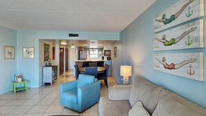Un salon avec un canapé bleu, un canapé beige et une petite table. Des tableaux encadrés ornent les murs, et une lampe est posée sur une table d’appoint.