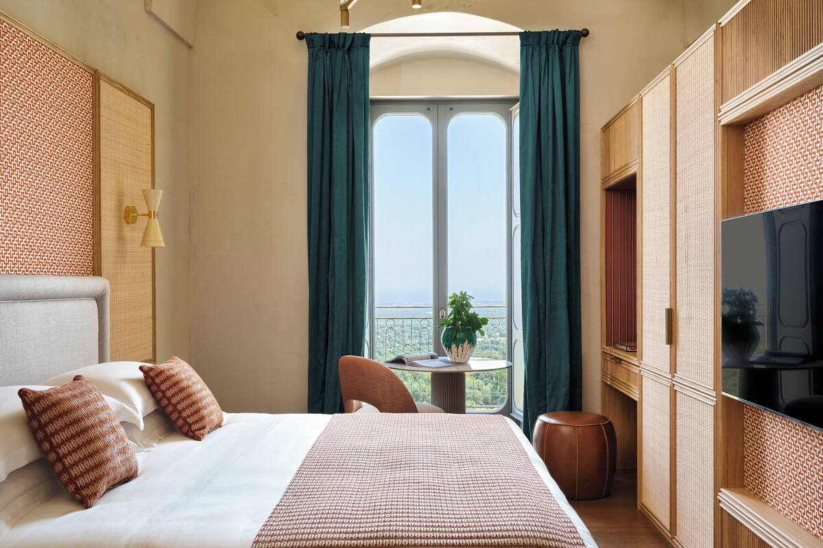 A hotel room with a large bed, a small table with a plant, a TV, and a balcony with a view.