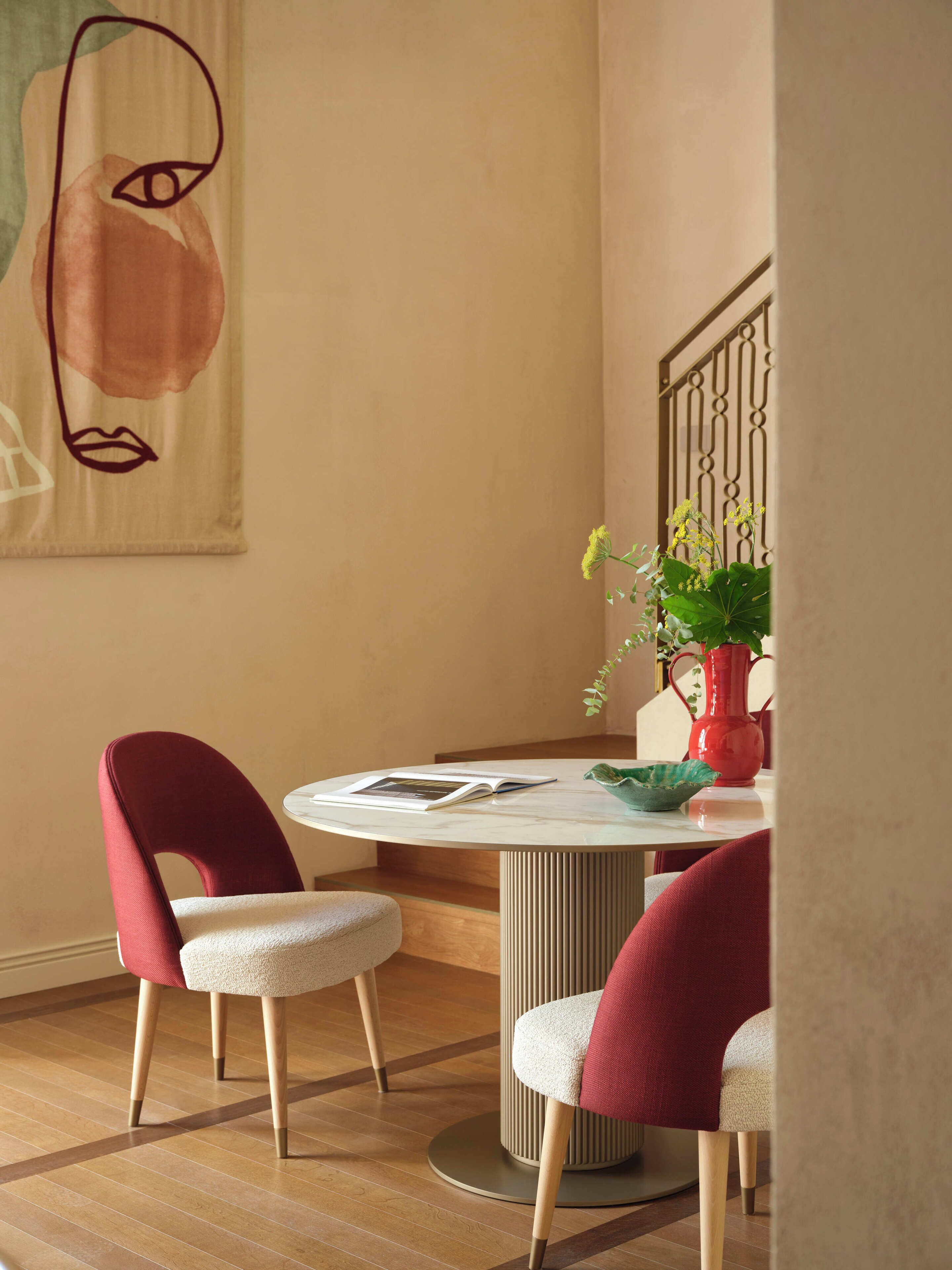 A dining area with a round table, two red upholstered chairs, a potted plant, and a wall-mounted abstract artwork.