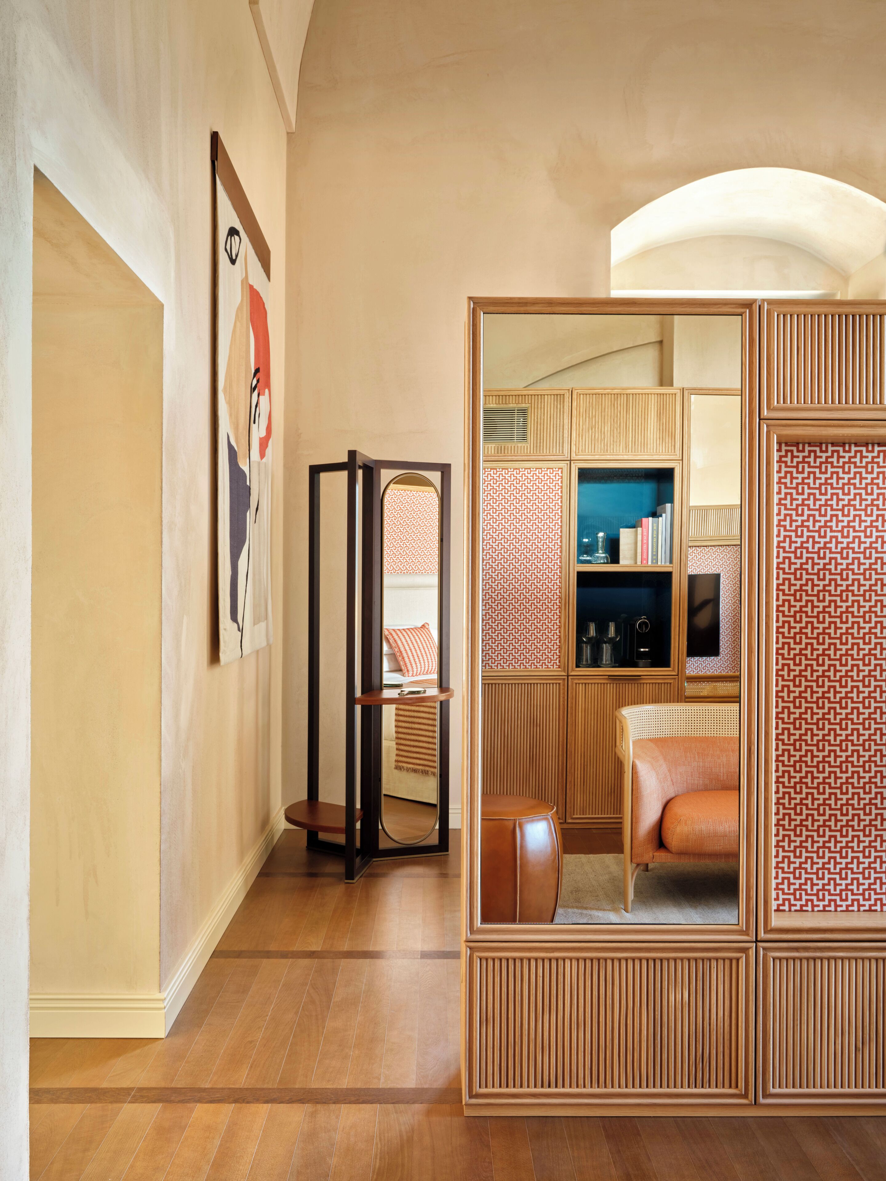 A modern hotel room with a wooden floor, a large mirror, a seating area with a patterned cushion, and a bookshelf.