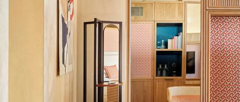 A modern hotel room with a wooden floor, a large mirror, a seating area with a patterned cushion, and a bookshelf.