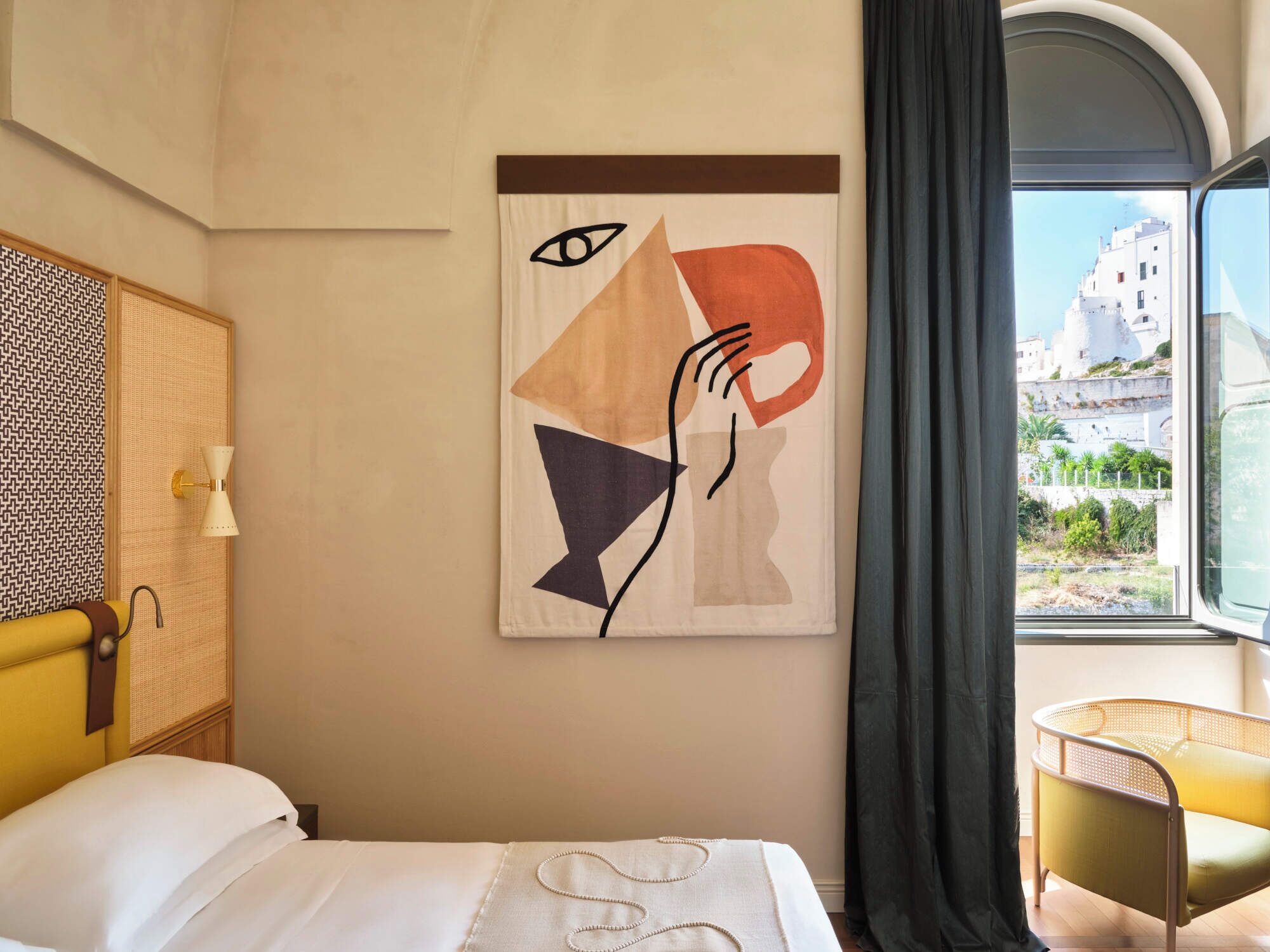 Une chambre d’hôtel avec un lit, une chaise, une œuvre d’art murale et une fenêtre donnant sur des immeubles.