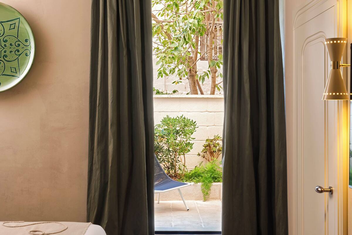 A hotel room with a bed, a view of a courtyard with plants, and a door with a light fixture.