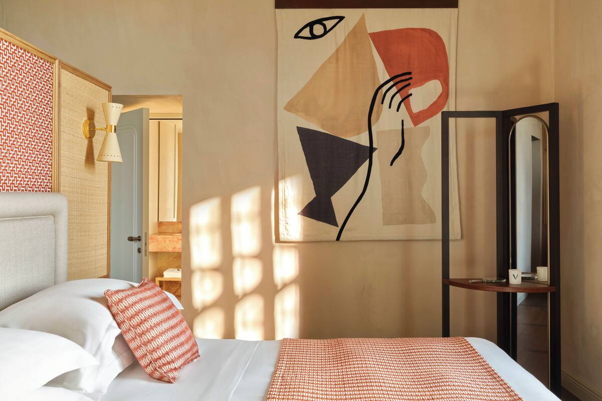 A hotel room with a bed, a wall-mounted abstract painting, a bedside table with a lamp, and a mirror.