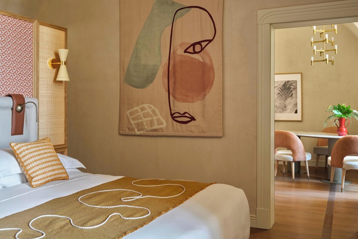 A hotel room with a large bed, a wall-mounted abstract painting, a dining area with chairs and a table, and a modern chandelier.