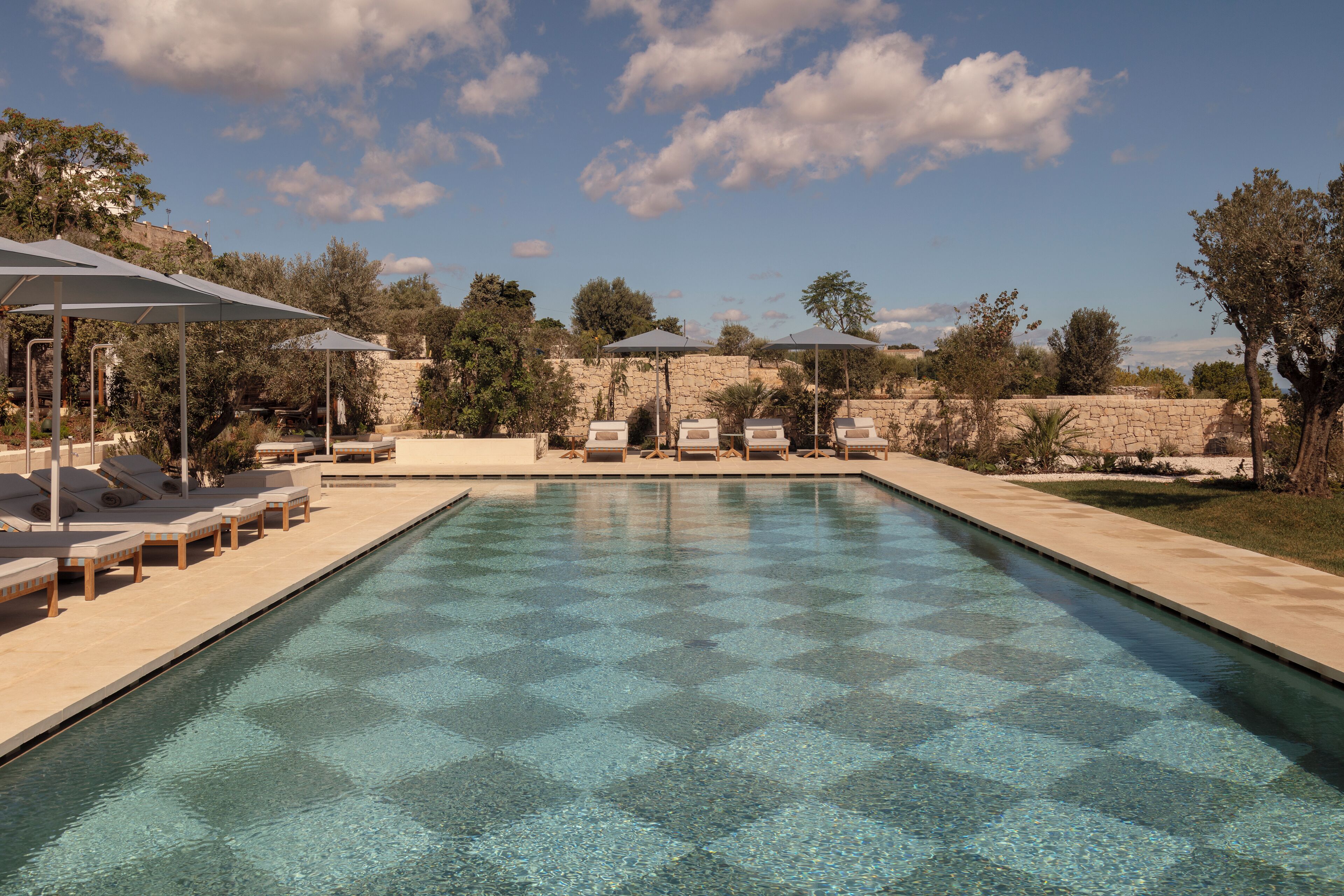 A swimming pool with lounge chairs and umbrellas, surrounded by stone walls and trees.