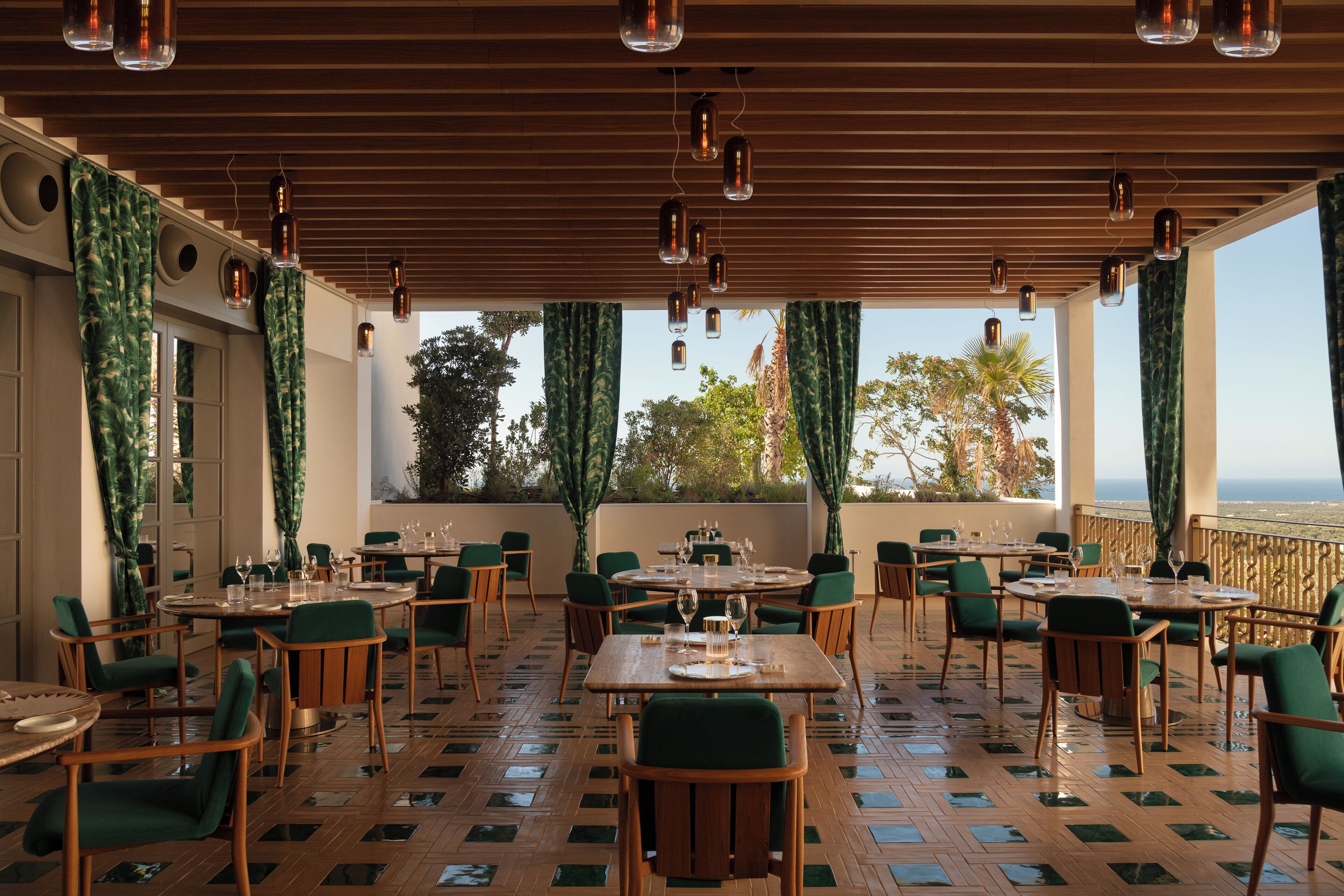 Ein Restaurant mit Holztischen und -stühlen, großen Fenstern mit Vorhängen und hängenden Pendelleuchten.