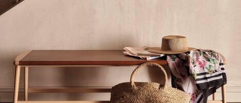 Un banco de madera con un bolso tejido y un sombrero frente a una pared con textura y una barandilla metálica.