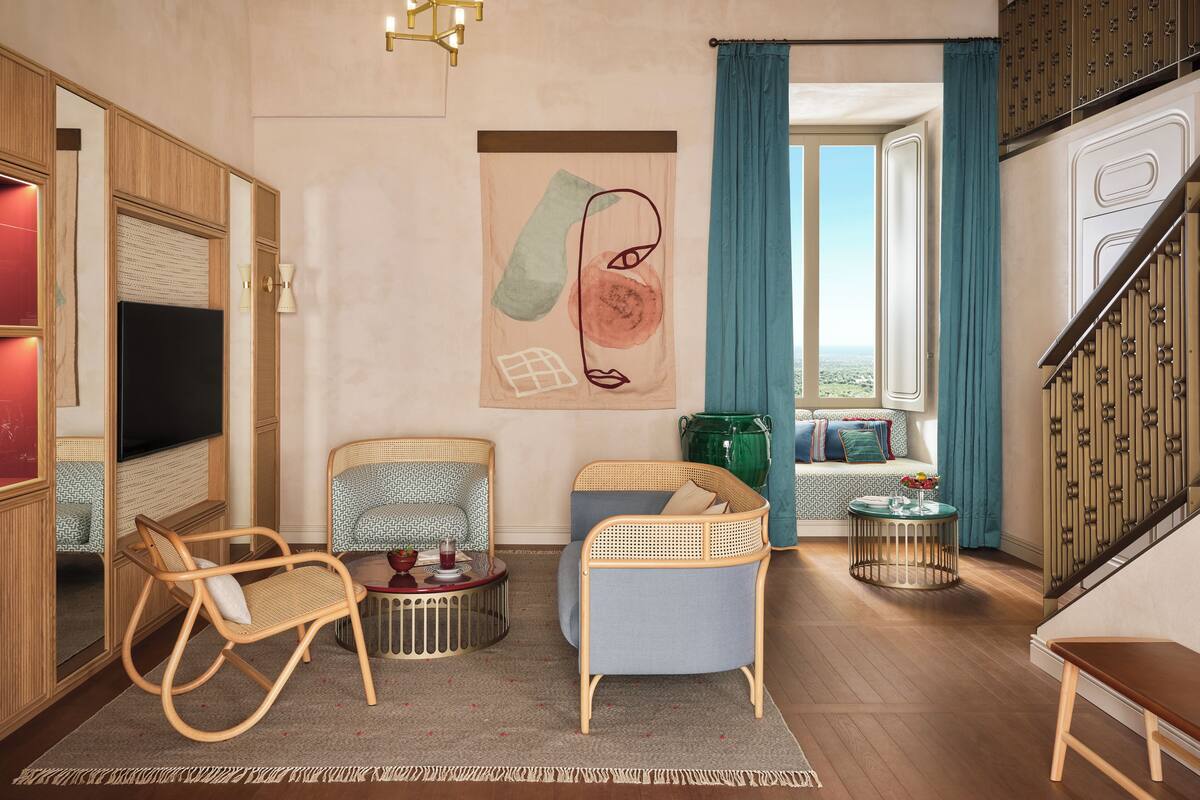 A modern living room with a flat-screen TV, wicker chairs, a round coffee table, and a view of the outdoors through a large window.