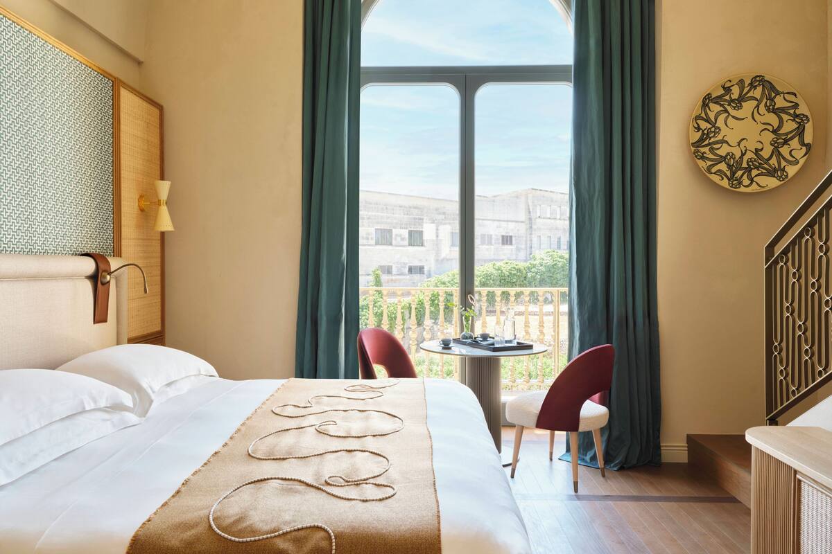 A hotel room with a large bed, a small table with chairs, a view of the city through a large window, and a decorative wall piece.