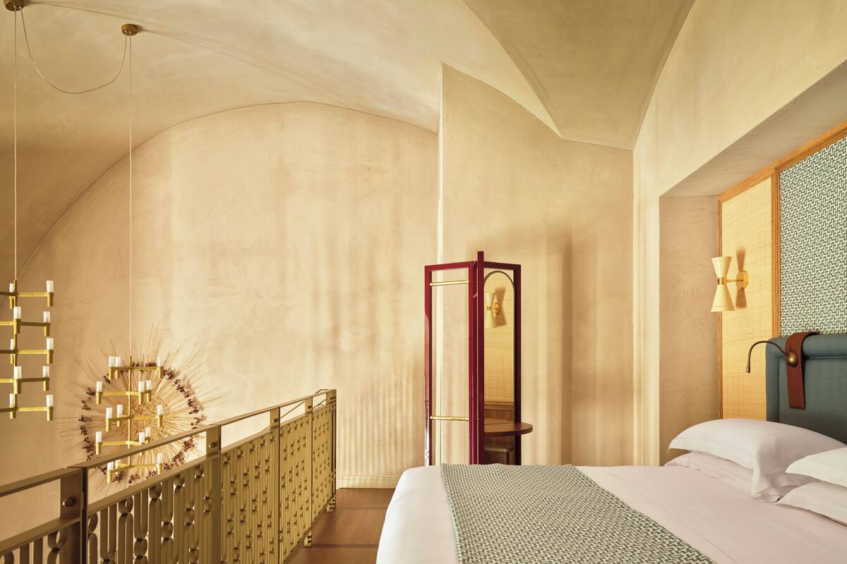 A hotel room with a curved ceiling, a bed with white linens, a wooden nightstand, a mirror, and a wall-mounted lamp.