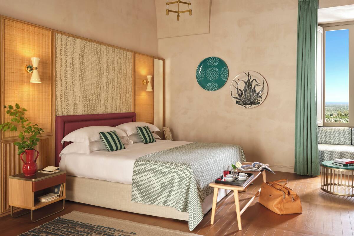 A modern hotel room with a large bed, bedside tables, a small table with a tray, a view of the city, and a balcony with a green curtain.