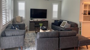 Een woonkamer met een flatscreen televisie, grijze banken, een bijzettafeltje en een plafondventilator.