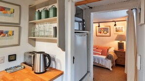 A compact kitchen with a toaster, kettle, and shelves holding glasses and green containers. A bed with a red and white striped blanket is visible in the adjacent room. - The Noel at Lesceave (Praa Sands)