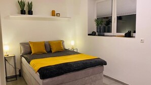 Un dormitorio ordenado con cama, lámparas de noche, un estante con plantas y una ventana con persianas.