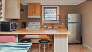 A compact kitchen with wooden cabinets, a stainless steel refrigerator, a microwave, a stove, and a sink. There are two stools under the counter. - ☀️2BR-Pet Friendly-1 Block from Beach-Off The Hook☀️ (St. Joe Beach)