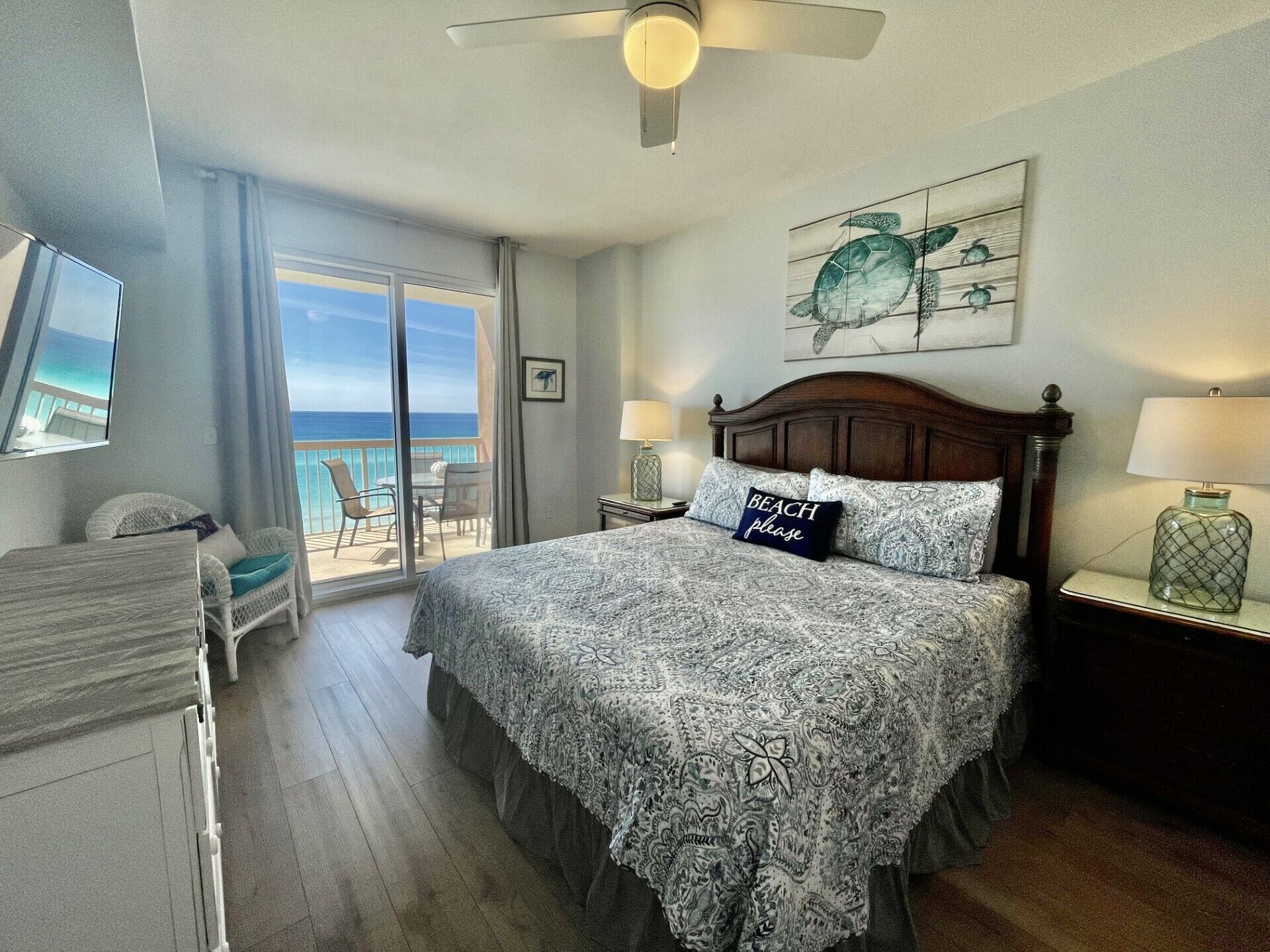 A bedroom with a large bed, a ceiling fan, a nightstand with a lamp, a chair, a window with a view of the ocean, and a painting of a turtle.