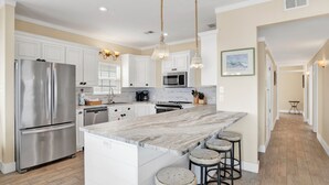 A modern kitchen with white cabinetry, stainless steel appliances, a marble island with bar stools, pendant lighting, and a view of the adjacent hallway. - Lagoon Front | Pet Friendly | Boat Launch, Pier and Beach Access (Gulf Shores)
