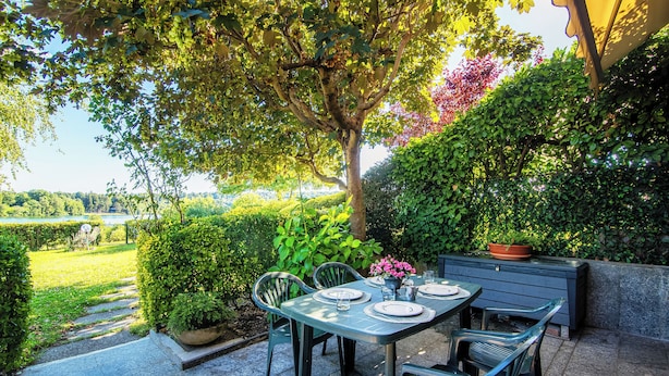 Un patio al aire libre con una mesa preparada para dos, rodeada de sillas, una planta en maceta y vistas a un jardín y un lago.