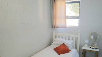 Cozy retreat with a single bed, nightstand, and fan, set against a minimalist backdrop of white walls and a tiled floor.